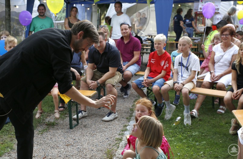 Ein Zauberer führt Kindern auf Bänken im Grünen einen Trick vor, viele Kinder sind gespannt und lachen.
