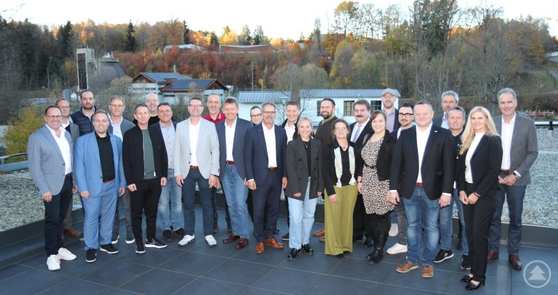 Die Unternehmerinnen und Unternehmer des IHK-Gremiums Freyung-Grafenau um ihren Vorsitzenden Jürgen Greipl (9.v.l.) sowie IHK-Hauptgeschäftsführer Alexander Schreiner (r.) diskutierten in den Wolfsteiner Werkstätten über die aktuellen Herausforderungen der regionalen Wirtschaft. Eine größere Gruppe von Männern und Frauen steht in mehreren Reihen auf einer Terrasse mit herbstlicher Landschaft im Hintergrund. Alle sind geschäftlich oder smart casual gekleidet und blicken lächelnd in die Kamera.