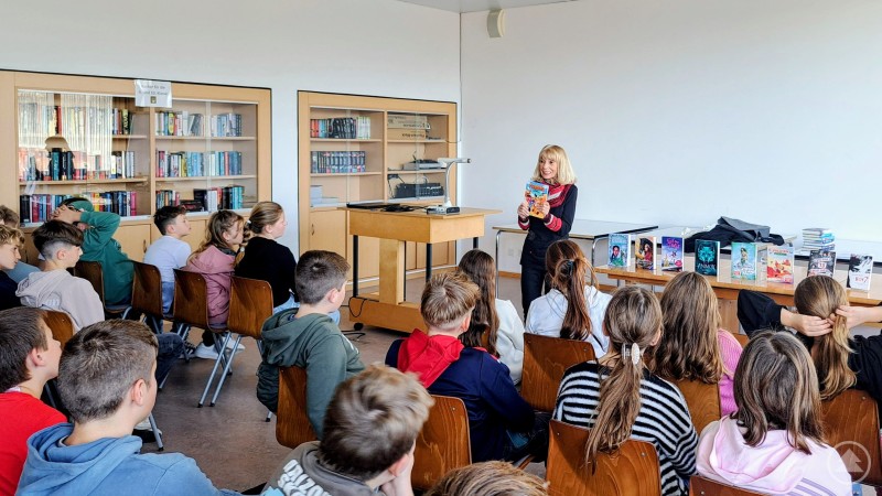 Hedwig Kunze von der Waldkirchener Buchhandlung präsentierte den 6.-Klässlern an der Realschule Grafenau eine bunte Mischung an Kinderbüchern. In einem hellen Klassenraum sitzen zahlreiche Schülerinnen und Schüler auf Stühlen und hören aufmerksam einer Frau zu, die ihnen ein Buch vorstellt. Im Hintergrund sind Regale voller Bücher und auf einem Tisch liegen verschiedene Kinder- und Jugendbücher aus.