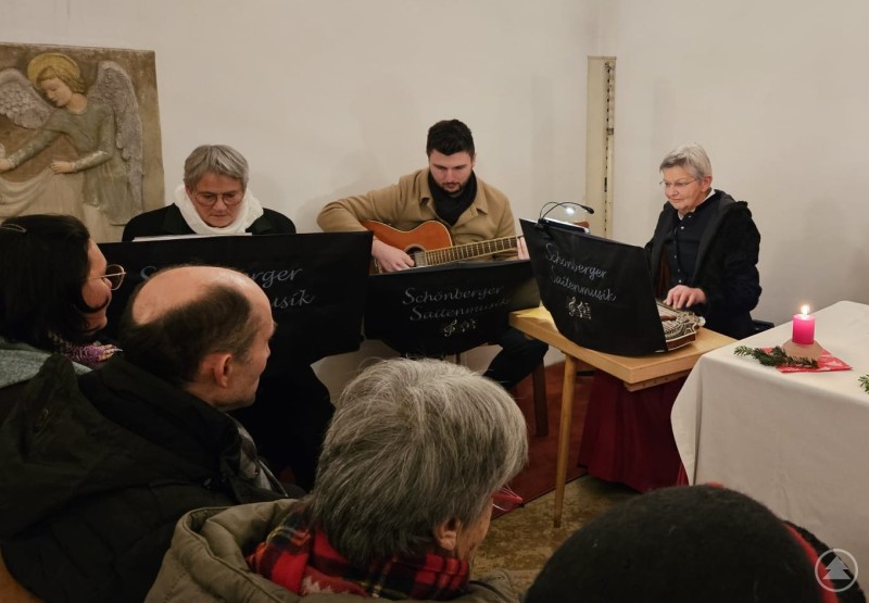 Die Schönberger Saitenmusi begleitete den Weg der Heiligen Familie nach Bethlehem, vorbei an bösen Wirtsleuten bis in den Stall mit Krippe, die Ochs und Esel trotz bayerischem Winter schon erwärmt hatten. Eine kleine Musikgruppe mit zwei Frauen und einem Mann spielt in einer Kirche traditionelle Instrumente, begleitet von Zuhörern. Auf den Notenständern steht "Schönberger Saitenmusik", neben der Gruppe brennt eine rote Kerze.