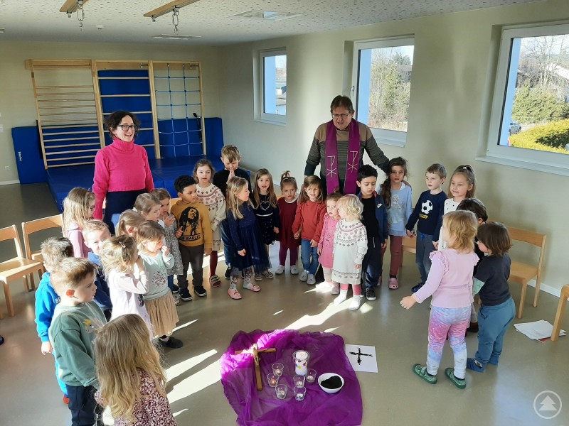 Pfarrer Andreas Artinger und Kita-Leitung Julia Greindl feiern gemeinsam mit den Kindern den Aschermittwoch im Kindergarten. Eine Gruppe Kindergartenkinder steht mit zwei Erwachsenen im Morgenkreis um ein mit einem violetten Tuch bedecktes Tuch, darauf liegen Kerzen und religiöse Symbole.
