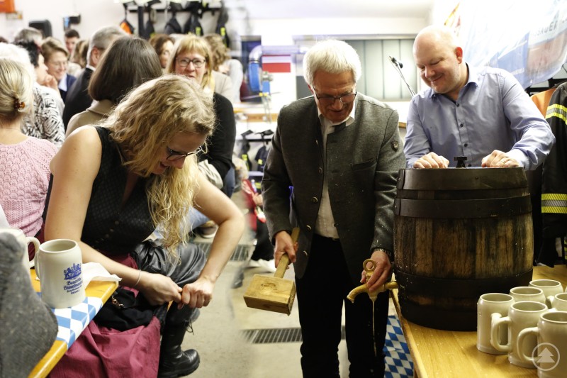 Mehrere Personen stehen in einem Feuerwehrhaus um ein Holzfass. Ein Mann schl&auml;gt mit einem Holzhammer auf den Zapfhahn, w&auml;hrend eine Frau daneben mithilft. Im Hintergrund sind G&auml;ste und Feuerwehruniformen zu sehen.