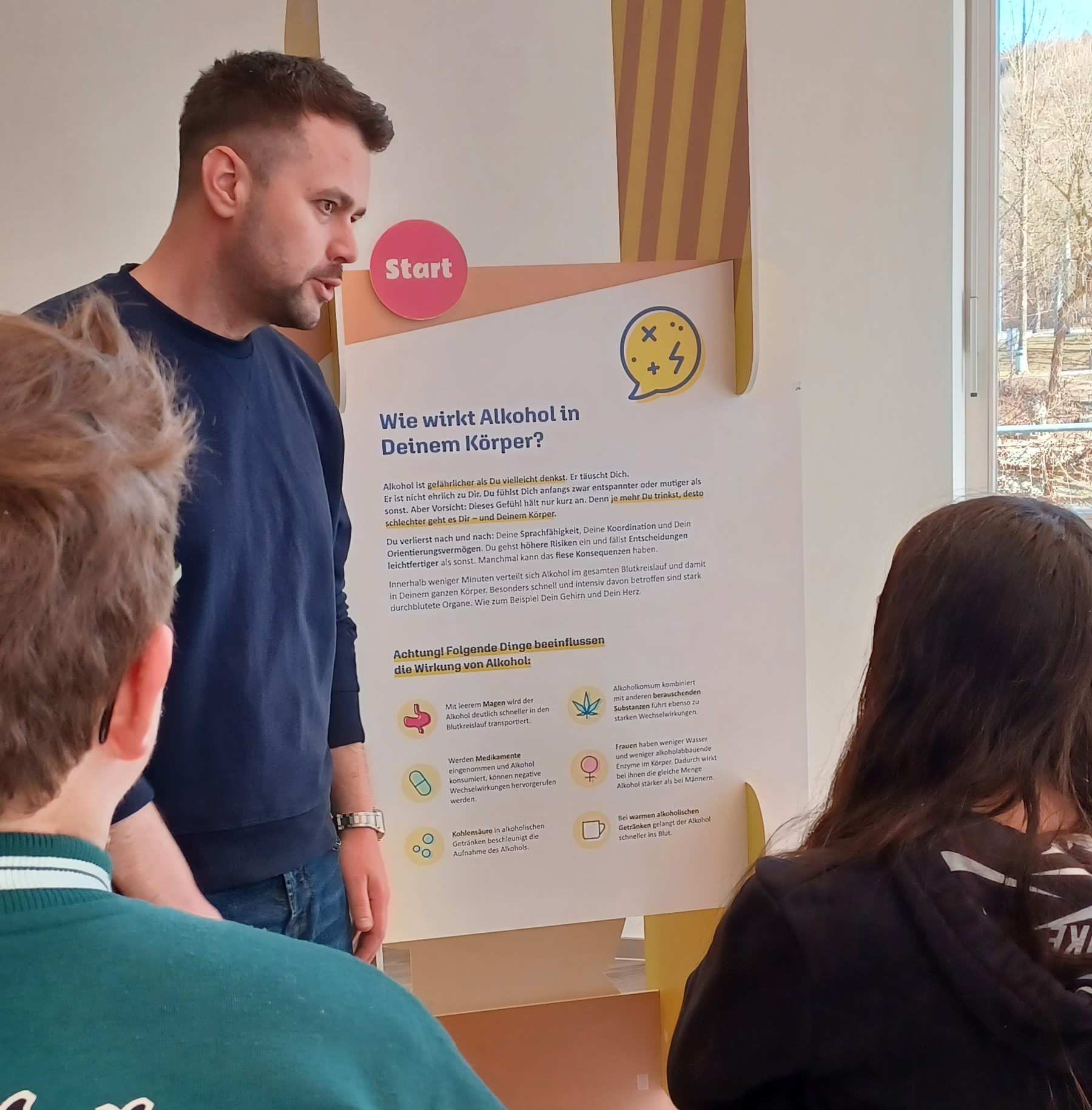 Auf dem Bild ist Christopher Kessel vom Gesundheitsamt Freyung-Grafenau zu sehen, wie er Jugendlichen im Rahmen der Ausstellung „Spaß ohne Punkt und Koma“ erklärt, wie Alkohol im Körper wirkt. Im Hintergrund steht ein großformatiges Infoposter mit dem Titel „Wie wirkt Alkohol in Deinem Körper?“, das anschaulich die körperlichen und psychischen Auswirkungen sowie beeinflussende Faktoren erläutert. Die Jugendlichen hören aufmerksam zu und nehmen die Informationen interessiert auf.