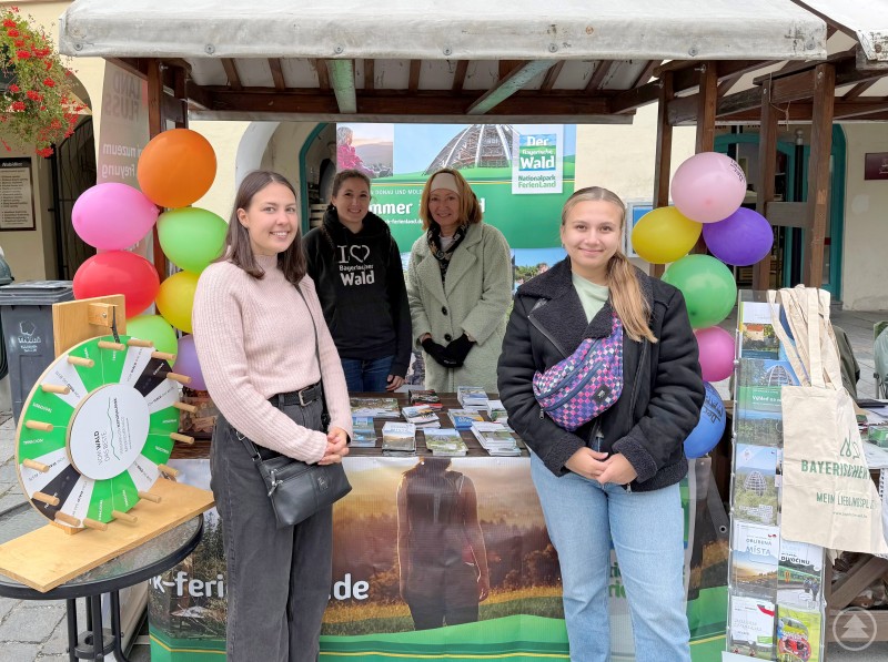 Gemeinsam präsentierten sie die Vielfalt des Landkreises Freyung-Grafenau und informierten die Besucherinnen und Besucher über touristische Angebote im Bayerischen Wald (von links) Praktikantin Jaroslava Pechová, Johanna Anetzberger, Selma Šedová-Keck (beide Landratsamt) sowie Praktikantin Alena Sigmundová. Vier Frauen stehen an einem bunt dekorierten Informationsstand mit Flyern und Glücksrad. Der Stand wirbt für den Bayerischen Wald und den Nationalpark.