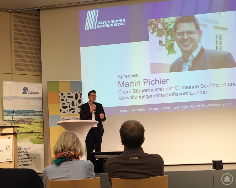 Bürgermeister Martin Pichler berichtete auf der Kommunale 25 in Nürnberg, wie Familienfreundlichkeit zur erfolgreichen Personalgewinnung in Kommunen beitragen kann. Martin Pichler, Erster Bürgermeister der Gemeinde Schönberg, spricht beim Bayerischen Gemeindetag am Rednerpult. Hinter ihm ist eine große Leinwand mit seinem Namen, Foto und Vortragstitel zu sehen.