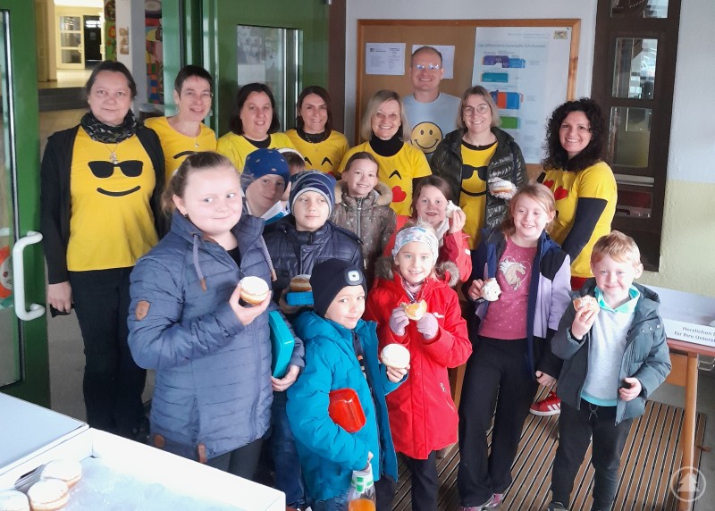 Mehrere Kinder und Lehrkräfte stehen in einem Schulgebäude zusammen. Einige Lehrkräfte tragen gelbe Shirts mit Smileys, die Kinder halten Krapfen in der Hand und lächeln in die Kamera.