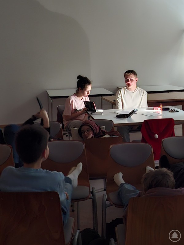 Spannende und schaurige Fälle der „drei Fragezeichen“ wurden an der Realschule Grafenau in abgedunkelter Atmosphäre von den Tutoren vorgelesen. Mehrere Kinder sitzen in einem abgedunkelten Klassenzimmer auf Stühlen und hören gespannt zu, während vorne zwei Jugendliche an einem Tisch mit Taschenlampe und Kerzenlicht Geschichten vorlesen.