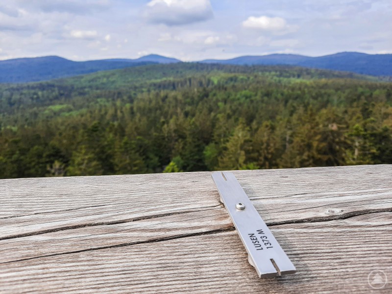Blick vom Baumwipfelpfad auf die sagenumwobene Landschaft zwischen Rachel und Lusen. Im Vordergrund ist eine hölzerne Brüstung mit einer Metallmarkierung zu sehen, auf der „Lusen 1373 m“ eingraviert ist. Dahinter breitet sich ein weiter, dichter Wald des Bayerischen Waldes aus. Am Horizont erheben sich sanfte, bewaldete Bergrücken unter einem teils bewölkten Himmel.