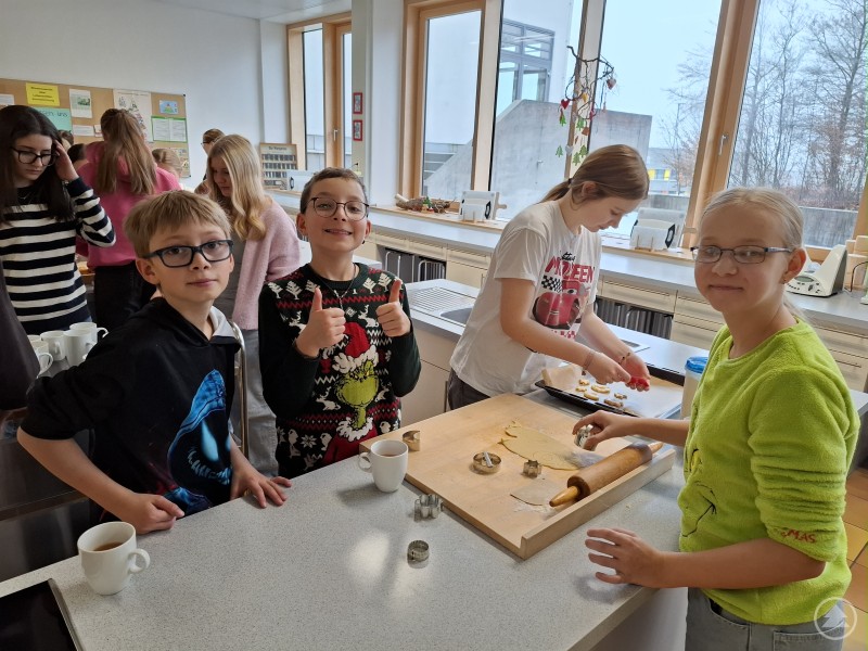 Die Grafenauer Realschüler aus den 5. Klassen backten am Adventsnachmittag begeistert Plätzchen. Mehrere Kinder stehen um eine Küchenzeile, stechen Plätzchen aus und lachen in die Kamera. Im Hintergrund wird an weiteren Tischen gebacken und gebastelt.