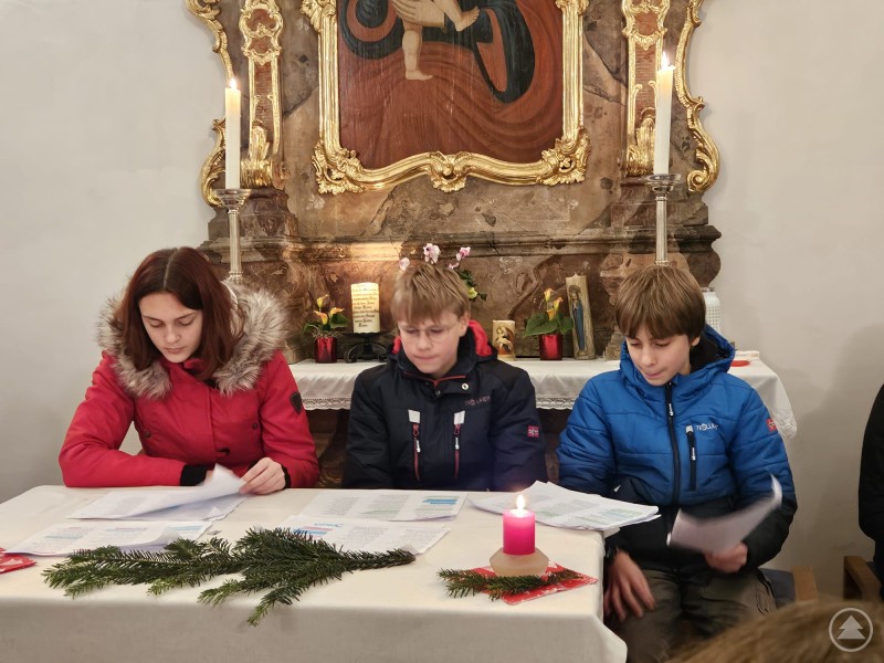 Drei Kinder sitzen an einem festlich geschm&uuml;ckten Tisch in einer Kirche. Vor ihnen liegen Manuskripte, Tannenzweige und eine rote Kerze. Im Hintergrund ist ein goldener Altar mit brennenden Kerzen zu sehen.