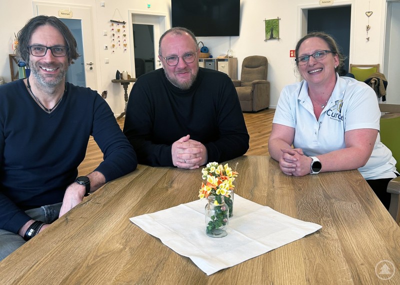 v.l.: Michael Schmid (Leitung Tagespflege Rosenium Freyung), Marco Binder (Leitung Tagespflege Caritas Waldkirchen und Freyung), Elisabeth Moser (Leitung Tagespflege Curare Waldkirchen) Drei Personen sitzen an einem großen Holztisch in einem hellen Gemeinschaftsraum und lächeln in die Kamera, auf dem Tisch steht eine kleine Blumenvase.