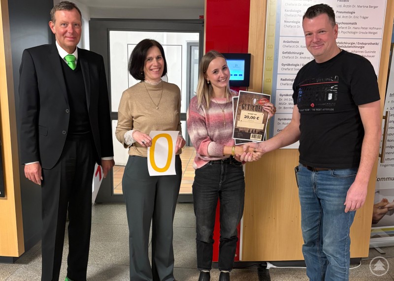Vier Personen stehen im Klinikgeb&auml;ude, eine Mitarbeiterin h&auml;lt einen Gutschein in der Hand, daneben stehen zwei Kolleginnen und ein Kollege.