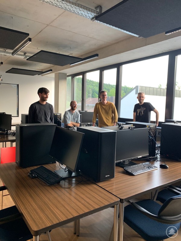 Teilnehmende des InnRaum³ Community-Treffens im INN.KUBATOR Passau beim Austausch in den Technikräumen. Vier junge Männer stehen in einem modernen, mit Computern ausgestatteten Raum und unterhalten sich. Im Vordergrund sind Monitore und Tastaturen auf Holztischen sichtbar.