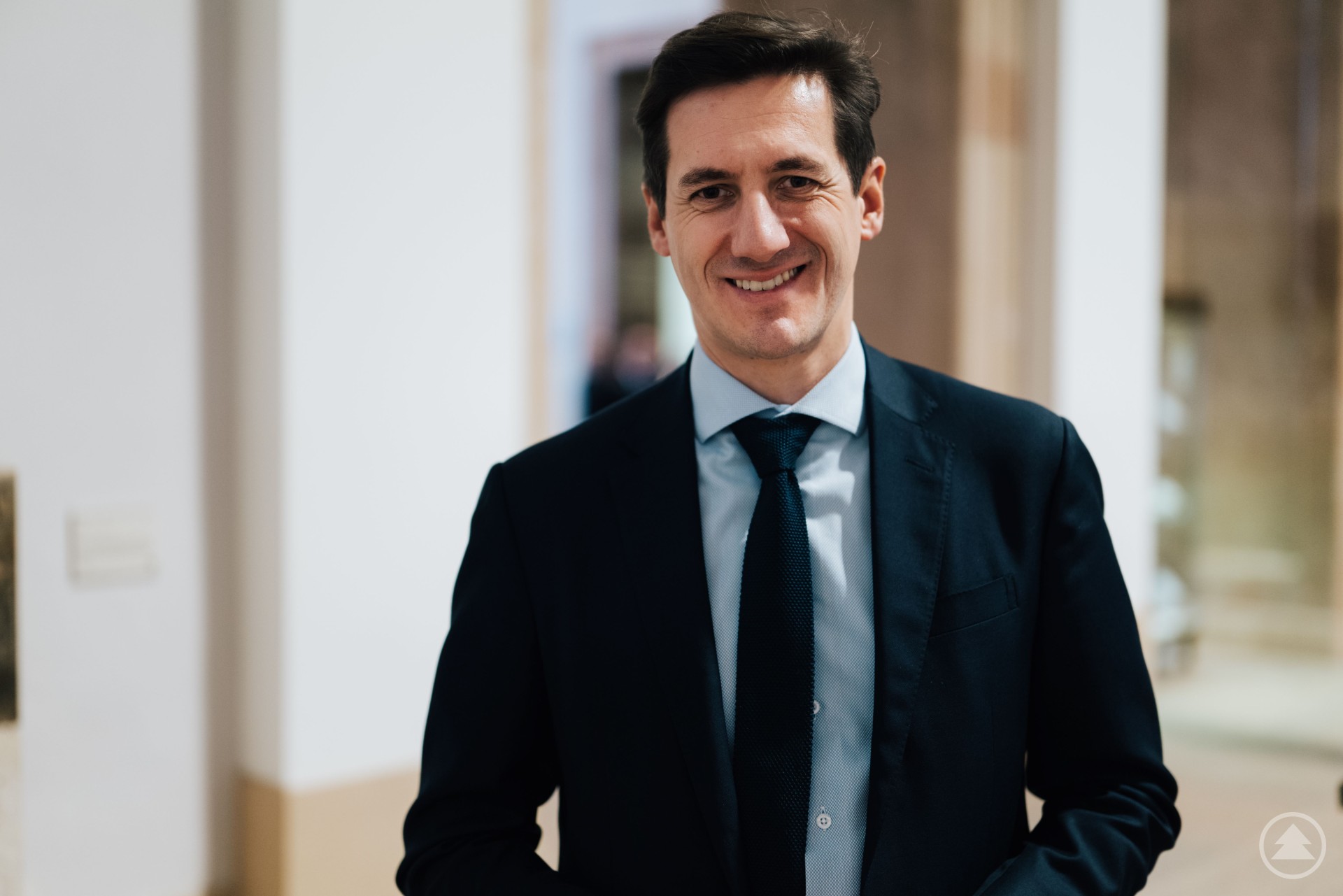 Ein Mann in Anzug und Krawatte steht lächelnd in einem hellen Flur mit weißen Wänden und Holztürrahmen.