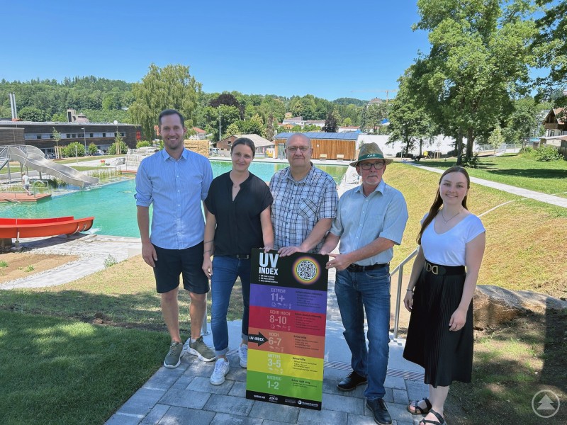 Auch das Naturfreibad in Freyung erhielt eine UV-Indextafel zur besseren Aufklärung der Badegäste. Fünf Personen präsentieren eine UV-Index-Tafel am Rand eines Naturfreibads mit Rutsche und Seeanlage.