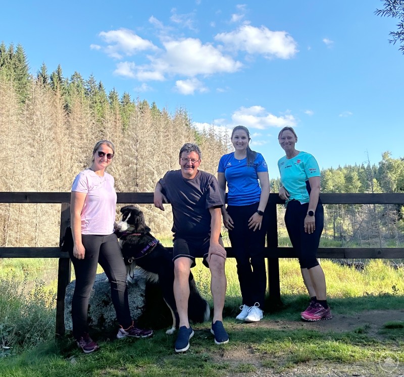 Konzeption des Questing-Wegs: Das Projektteam (von links) Chiara Löw, Martin Zellner, Johanna Anetzberger und Claudia Schmid arbeitet im Anschluss an die Begehung intensiv an der inhaltlichen Ausarbeitung der Fragen für den neuen Questing-Weg in Bischofsreut. Vier Personen und ein Hund stehen bei Sonnenschein an einem Holzzaun mit Blick auf den Wald, sie lächeln in die Kamera.