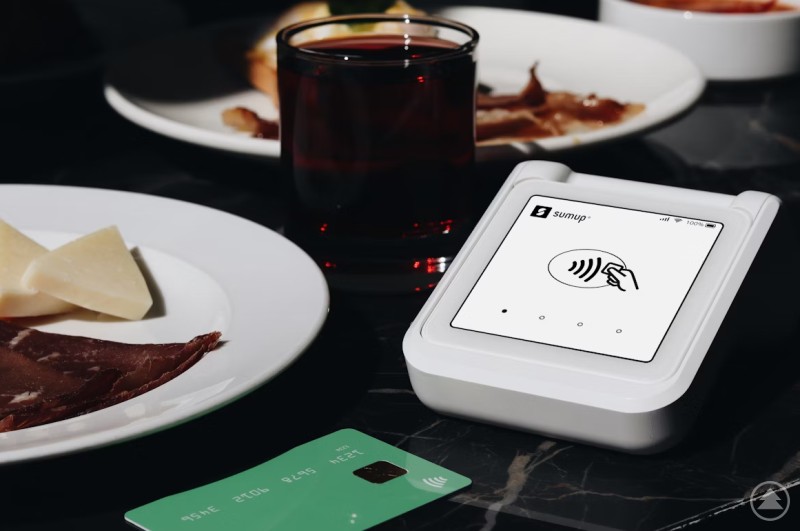 ChatGPT: Auf dem Bild ist ein moderner, weißer Kartenleser von SumUp zu sehen, der für kontaktlose Zahlungen bereit ist. Daneben liegen eine grüne Kreditkarte, ein Glas mit einem dunklen Getränk (vermutlich Rotwein) sowie ein Teller mit Käsestücken und Schinken. Die Szene wirkt wie ein gedeckter Restauranttisch mit Fokus auf das bargeldlose Bezahlen.