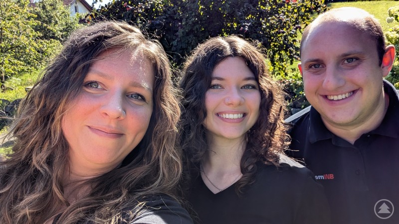 Kathleen (Kathi) Müller, Jana Spannbauer und Andreas Bildl vom BWmedien-Team glauben fest an die digitale Kraft regionaler Gemeinschaft. Drei Personen stehen lächelnd im Grünen, bei Sonnenschein, im Hintergrund sind Bäume und ein Haus zu sehen.