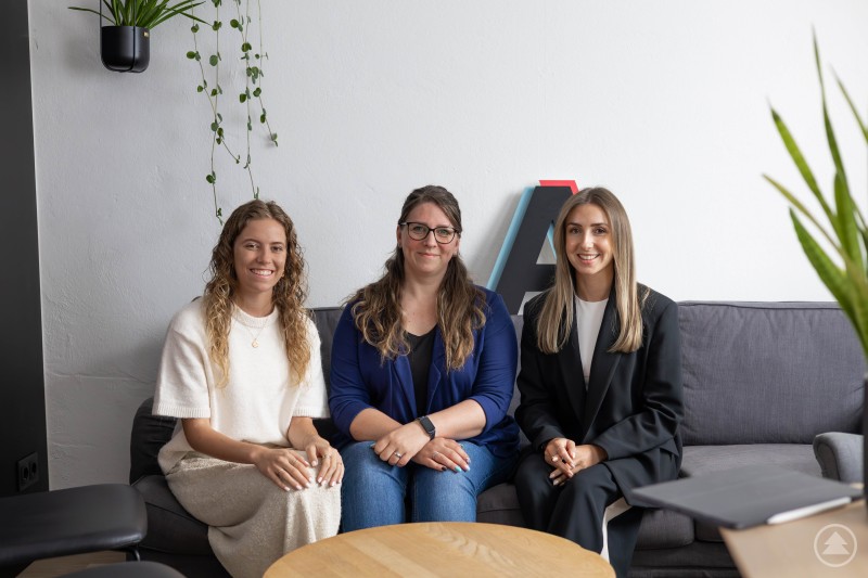 Kathi Lenz, Kathleen Müller und Eva Nirschl beim gemeinsamen Talk im AzubiTok-Studio – offen, herzlich und ganz nah dran an Kathleens beruflichem Neustart. Drei Frauen sitzen gemeinsam auf einem Sofa in einem modern eingerichteten Büro oder Besprechungsraum. Sie lächeln in die Kamera, wirken freundlich und professionell. Links hängt eine Pflanze an der Wand, rechts und vorne sind weitere Pflanzen zu sehen. Hinter ihnen steht ein großes Deko-Element mit farbigen Kanten.