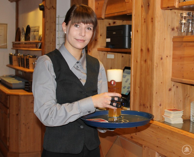 Nicole Kufner bei ihrer Arbeit als Restaurantfachfrau. Eine Restaurantfachfrau mit dunklem Pony und Dienstkleidung serviert ein frisch gezapftes Bier auf einem Tablett. Sie trägt an der rechten Hand eine spezielle Teilfingerprothese, die ihr das Arbeiten trotz Handicap ermöglicht. Die Szene spielt in einem gemütlichen, holzgetäfelten Gastraum.
