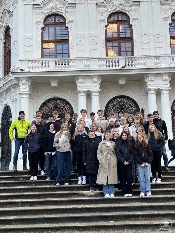 Eindrucksvolle Momente genossen die Grafenauer Realschüler der Klassen 10b und 10c bei ihrer Abschlussfahrt in der österreichischen Hauptstadt. Eine große Gruppe von Schülerinnen, Schülern und Lehrkräften steht auf einer Treppe vor einer prunkvollen, historischen Fassade mit weißen Säulen und reich verzierten Fenstern. Alle blicken lächelnd in die Kamera und sind winterlich gekleidet.