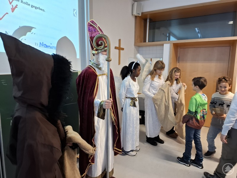 Der Nikolaus steht mit Bischofsstab und Mitra neben Engeln und einem Krampus in einem Klassenzimmer. Kinder stehen in einer Reihe und bekommen Geschenke &uuml;berreicht.