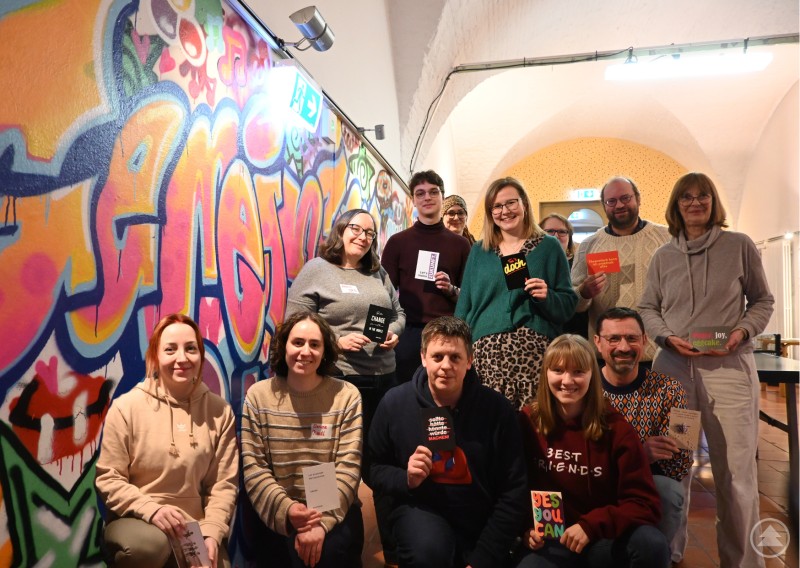 Teilnehmer des Fachtages im Jugendzentrum Zeughaus Eine Gruppe von Erwachsenen posiert in einem Raum vor einer bunten Graffiti-Wand und hält Unterlagen in die Kamera.