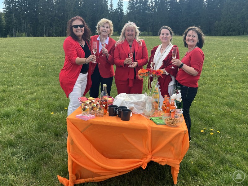 Fünf Frauen in roten Outfits stoßen mit Sektgläsern an einem dekorierten Tisch in einer Wiese an. Im Hintergrund sind Bäume zu sehen.