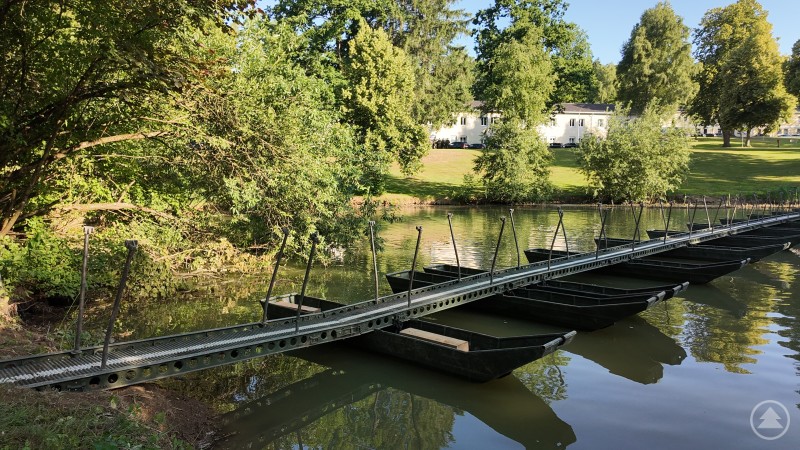 Ein Pontonsteg aus mehreren Metallbooten überquert einen ruhigen Fluss, eingebettet in eine grüne Parklandschaft.