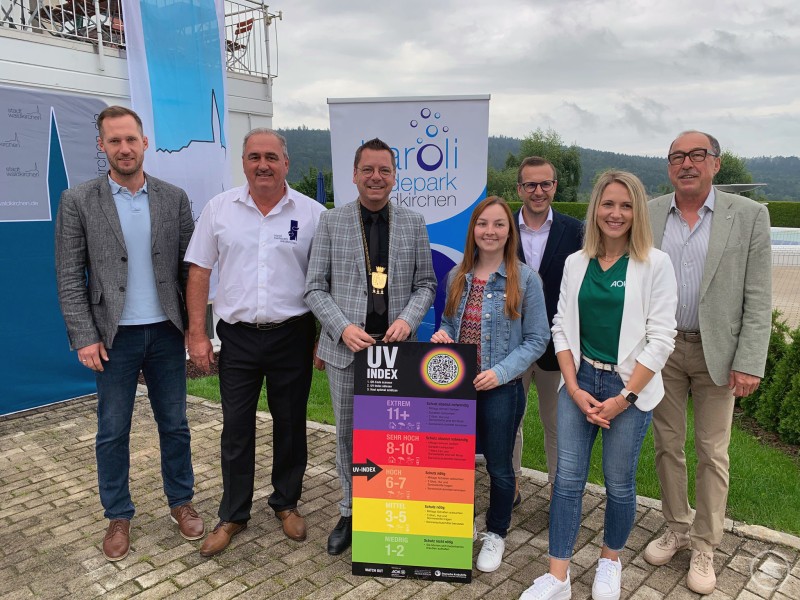 Übergabe der UV-Indextafel im Karoli-Badepark Waldkirchen: Vertreter der Stadt, des Landkreises, der AOK und der Gesundheitsregion Plus setzen ein Zeichen für Sonnenschutz. Gruppe von sieben Personen steht vor Bannern und einer UV-Index-Tafel, im Hintergrund der Außenbereich eines Schwimmbads.