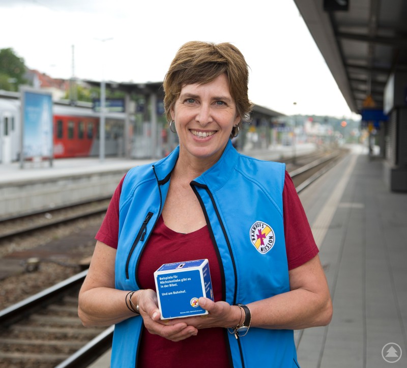 Ein sicherer Hafen in Passau: Angelika Leitl-Weber und ihr Team in der Bahnhofsmission helfen Menschen ohne Unterkunft. Eine Frau in rotem Shirt und blauer Weste steht lächelnd auf einem Bahnsteig und hält eine kleine Box in den Händen. Im Hintergrund sind Gleise und ein Zug zu sehen.