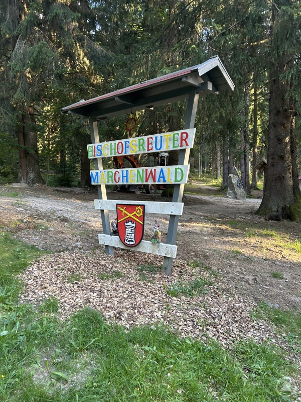 Start- und Zielpunkt des Questing-Wegs: Der Bischofsreuter Märchenwald lädt nicht nur zum Staunen ein, sondern bildet auch den Ausgangspunkt für die spannende Rätseltour durch den Ort. Ein hölzernes Schild mit der bunten Aufschrift „Bischofsreuter Märchenwald“ steht zwischen hohen Bäumen im Wald.