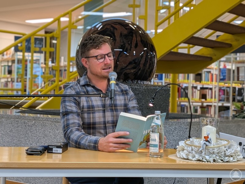 Leo Reisinger bei seiner Lesung in der Kreisbibliothek Freyung: Mit Auszügen aus seinem Debütroman „Bavarese“ begeisterte der Schauspieler das Publikum. Leo Reisinger steht mit Mikrofon in der Hand vor Publikum in der Kreisbibliothek Freyung. Hinter ihm sind Bücherregale und die markante gelbe Wendeltreppe zu sehen.