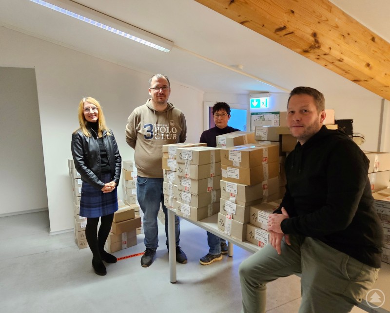 v.l.: Karin Schlappinger (Abteilung Kommunikation), Oliver Pöllendorfer (Mitarbeiter WiP), Barbara Dichtl (Gruppenleiterin WiP), Andreas Schurm (Technischer Leiter) Vier Personen stehen und sitzen in einem hellen Raum an einem Tisch, der mit vielen gestapelten Versandkartons gefüllt ist. Die Atmosphäre wirkt freundlich und kollegial, im Hintergrund sind Dachbalken und eine Notausgangsbeschilderung zu sehen.