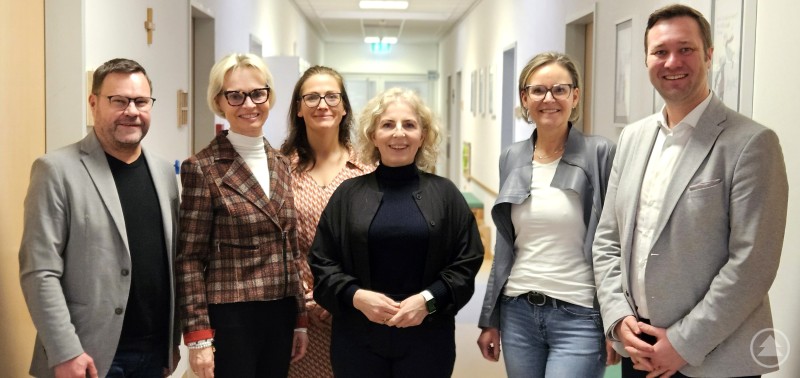 Nahaufnahme eines Gruppenfotos: Sechs Personen stehen in einem Flur einer Einrichtung, freundlich zur Kamera gewandt. Der Flur ist hell beleuchtet, an den W&auml;nden h&auml;ngen Bilder.