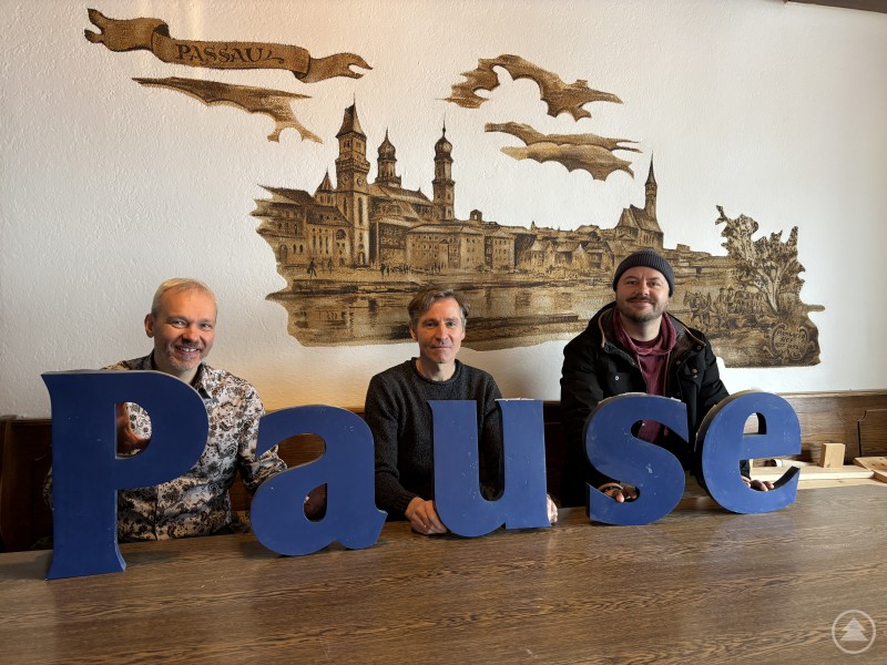 Der ehemalige Stammtisch im Passauer Hof wird wiederbelebt – die Pause ist vorbei. Libor Pražan, Thomas Petermaier und Uli Hansel haben schon Probe gesessen v.li. Drei Männer sitzen an einem großen Holztisch im ehemaligen Gastraum und halten blaue Buchstaben, die zusammen das Wort Pause ergeben.