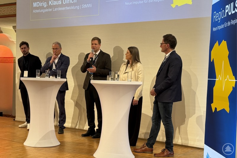 Bezirkstagspräsident Dr. Olaf Heinrich (Mitte) diskutierte mit Julian Menninger (von links), MDirig Klaus Ulrich, Anja Zöllner-Sprotte und Prof. Dr. Markus Lemberger auf dem Podium der Veranstaltungsreihe Regio.Puls über Antworten auf regionale Herausforderungen. Podiumsdiskussion mit fünf Personen auf einer Bühne, ein Redner mit Mikrofon steht in der Mitte, daneben weitere Teilnehmer an Stehtischen, im Hintergrund eine Präsentationswand.