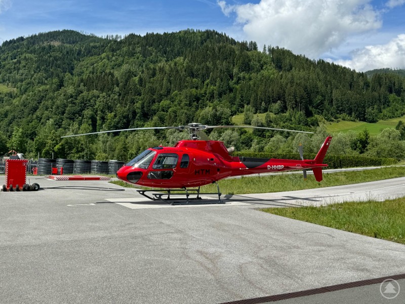 Dieser Hubschrauber hebt in Aidenbach ab: AS350, eine Single-Engine-Maschine mit Platz für bis zu fünf Passagiere. Roter Hubschrauber vom Typ AS350 am Boden mit Wald- und Bergkulisse im Hintergrund.