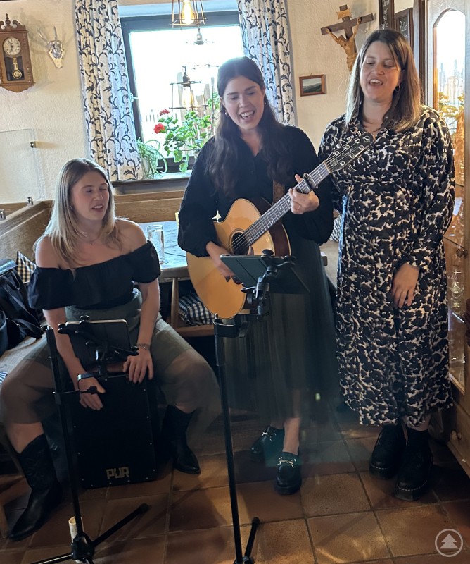 Die Lieblings-Damenband von Roswitha Prasser, das Geschwistertrio „eXCLent“, war eine emotionale Überraschung für die ehemalige Vereinsvorsitzende. Drei junge Frauen musizieren in einer urigen Gaststube – eine spielt Gitarre, eine sitzt am Cajón, die dritte singt. Alle wirken emotional und engagiert.