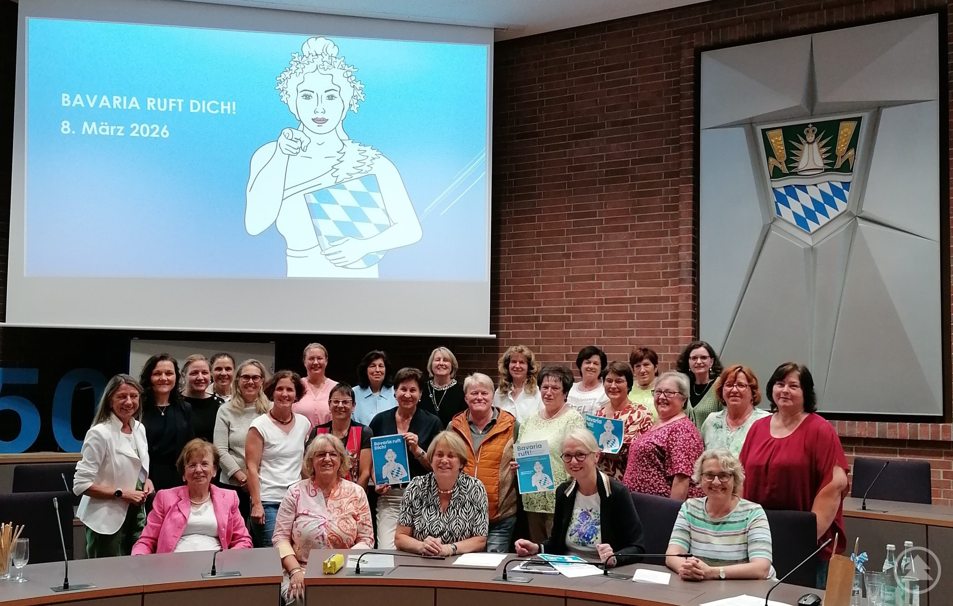 Die Kommunalpolitikerinnen beim Gruppenfoto im Großen Sitzungsaal. Gruppe von etwa 30 Frauen unterschiedlichen Alters posiert gemeinsam im großen Sitzungssaal eines Landratsamts, im Hintergrund ein Bildschirm mit dem Schriftzug „BAVARIA RUFT DICH – 8. März 2026“ und eine Illustration einer Frau in bayerischer Tracht.