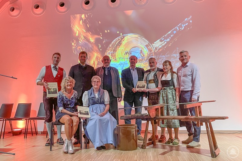 Präsentierten das neue Buch: Martin Wolf (hinten v. l.), Dr. Olaf Heinrich, Michael Haug, Dr. Andreas Bernhard, Karl Dudek, Marianne Wolf, Alfons Schinabeck, Hilde Greiner (vorne v. l.) und Paula Dudek. Neun Personen stehen und sitzen auf einer Bühne vor einer leuchtenden Projektionswand mit einem stilisierten Glasmotiv. Einige von ihnen halten das vorgestellte Buch „Der weite Weg des Glases“ in den Händen.