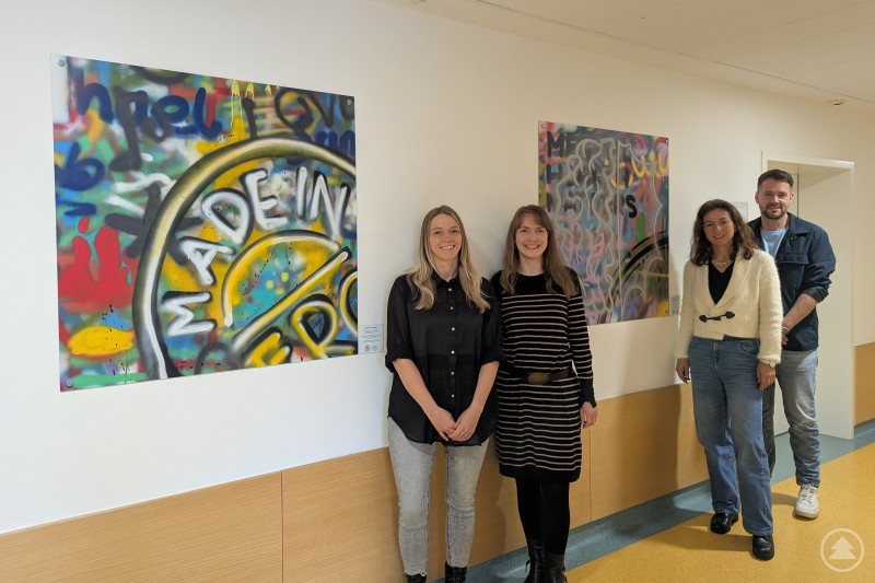 Vier Erwachsene stehen lächelnd in einem hellen Flur vor zwei bunt gesprayten Kunstwerken, die nun im Gesundheitsamt ausgestellt sind.