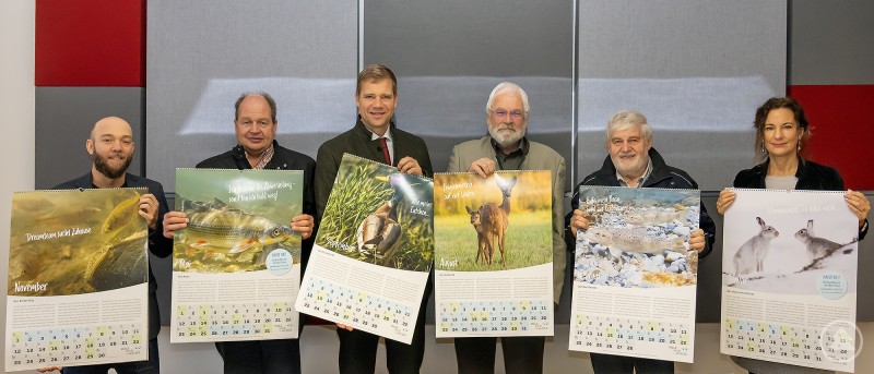 Im Bild (v.l.): Fabio di Tullio, Michael Kreiner, Bezirkstagspräsident Dr. Olaf Heinrich, Christoph Dusch, Herbert Mayer und Nicola Holzapfel Sechs Personen stehen nebeneinander und halten jeweils einen großen Wandkalender mit Tiermotiven in den Händen. Auf den Kalenderblättern sind verschiedene Wildtiere und Gewässer abgebildet. Im Hintergrund ist eine moderne Wand mit farbigen Akustik-Paneelen zu sehen. Alle Personen blicken freundlich in die Kamera.