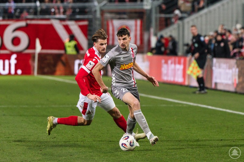 Ein Spieler des SSV Jahn Regensburg im grauen Trikot setzt sich an der Seitenlinie gegen einen Gegenspieler in Rot durch. Im Hintergrund Zuschauer und Linienrichter.
