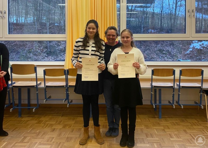 Die Blockflötistinnen Louisa Geißdörfer (vorne, v. l.) und Sophie Schwemmer mit Lehrerin Inge Reinelt. Zwei Mädchen präsentieren stolz ihre Urkunden, begleitet von ihrer Lehrerin in einem Klassenraum mit Stühlen im Hintergrund.