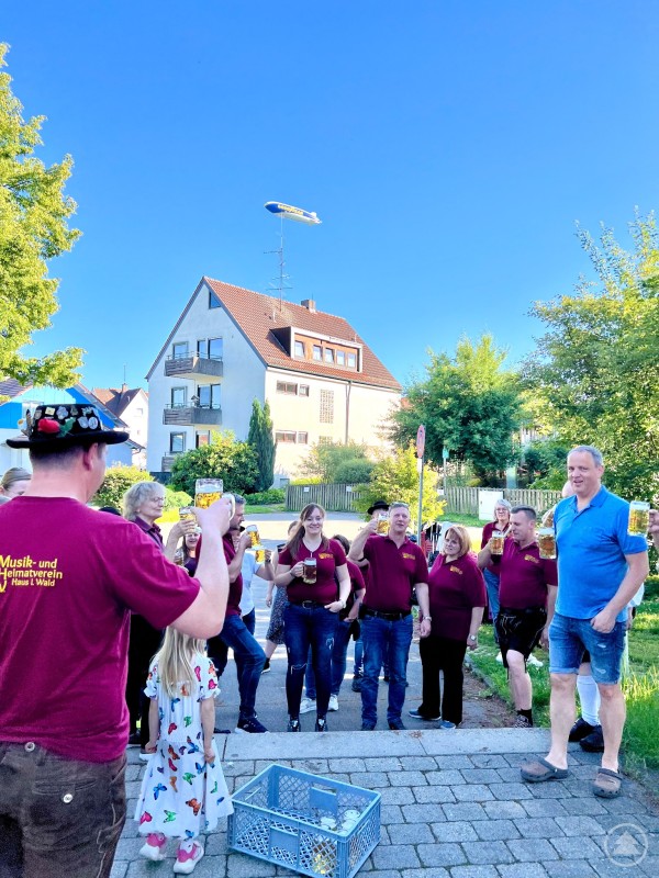 Eine Gruppe von Menschen in roten Vereinsshirts stößt mit Maßkrügen im Freien an, im Hintergrund fliegt ein Zeppelin über ein Wohnhaus.