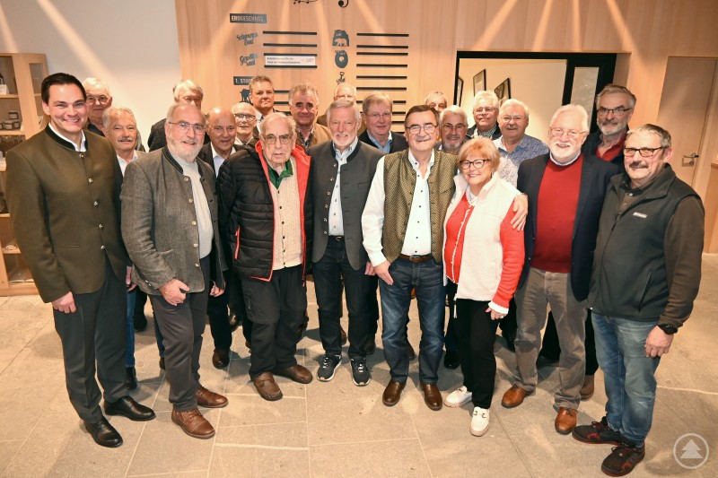 Lebhafte Gespräche – über frühere Amtszeiten, besondere Erinnerungen, aktuelle Entwicklungen in den Gemeinden und natürlich auch zu den bevorstehenden Kommunalwahlen prägten das Treffen der ehemaligen Bürgermeister in der Volksmusikakademie. Eingeladen dazu hatten Landrat Sebastian Gruber (links) und der Kreisvorsitzende des Bayerischen Gemeindetags, Bürgermeister Josef Kern (9. von rechts). Eine größere Gruppe älterer Männer und Frauen steht fröhlich zusammen in einem modernen Innenraum. Im Hintergrund sind Wandbeschriftungen, Holzwände und Ausstellungselemente zu erkennen. Die meisten Personen tragen Trachten- oder Freizeitkleidung, einige umarmen sich oder lächeln in die Kamera.