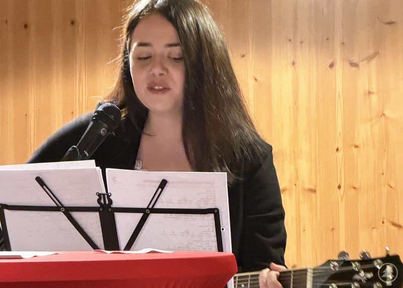 Eine Frau steht an einem Rednerpult und singt mit Gitarre vor Holzvert&auml;felung. Vor ihr befindet sich ein Notenst&auml;nder mit Liedtexten.