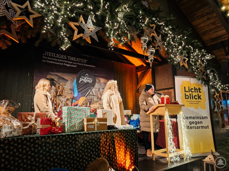 Die beiden Päckchenversteigerungen erzielten über 4000 Euro für die Lichtblick Seniorenhilfe, v.l. Lisa Peter vom Stadtmarketing, Weihnachtsengel Bianca, Konrad Wilhelm von Lichtblick. Drei Personen stehen auf einer weihnachtlich geschmückten Bühne, die mit Lichterketten und Sternen dekoriert ist. Eine Frau spricht am Rednerpult ins Mikrofon, während vor ihr mehrere bunte, fest verpackte Päckchen stehen. Im Hintergrund ist ein Banner von Lichtblick Seniorenhilfe zu sehen, das auf den Einsatz gegen Altersarmut hinweist. Die Atmosphäre ist festlich und lebendig.