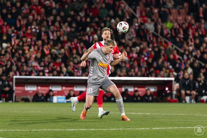 Harter Luftkampf: Die Jahnelf behauptet sich auch in der Defensive gegen den Tabellenführer. Ein Spieler des SSV Jahn Regensburg im grauen Trikot köpft den Ball unter Bedrängnis von einem Cottbus-Spieler. Im Hintergrund ist die mit Fans gefüllte Tribüne zu sehen.